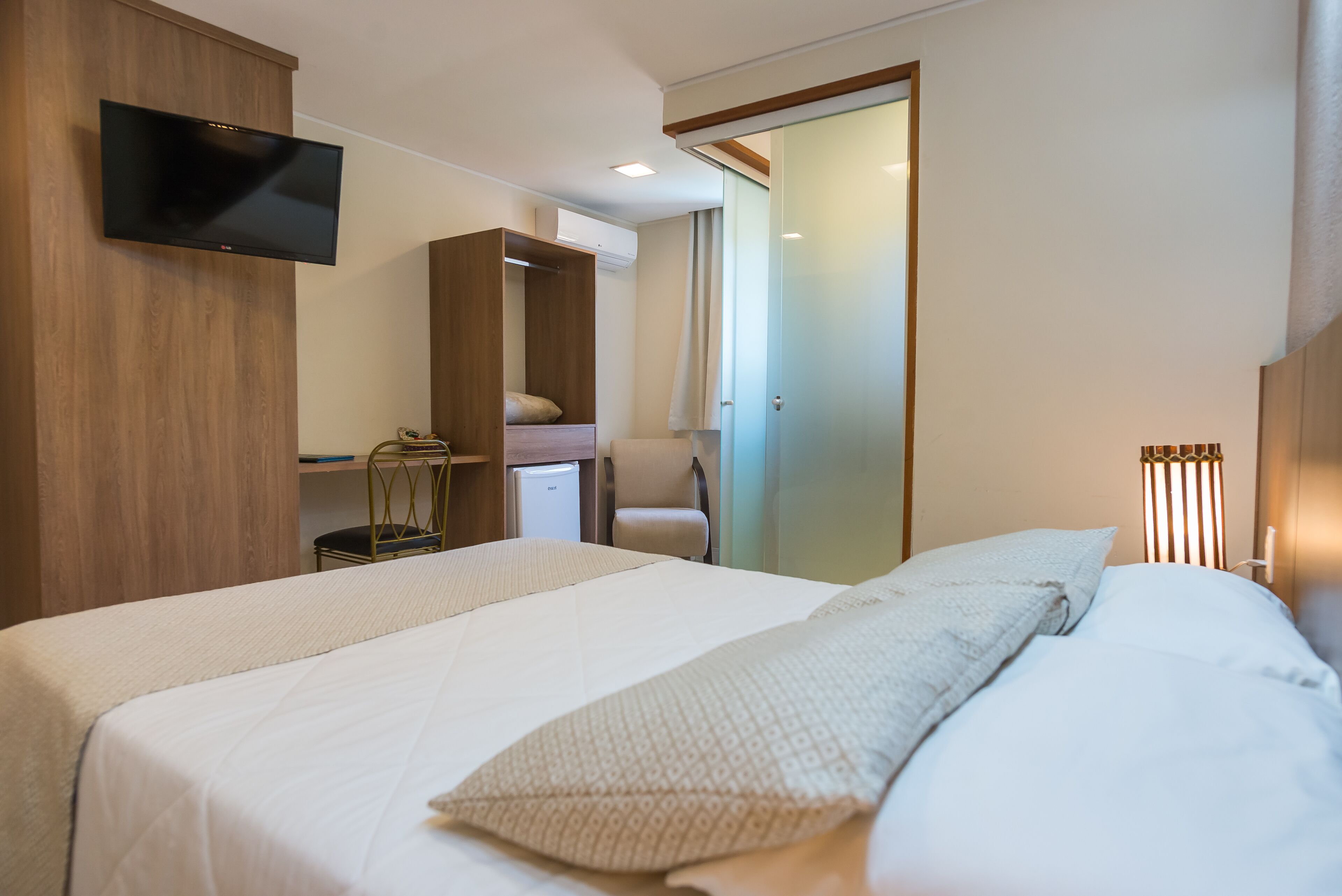Ein Hotelzimmer mit einem großen Bett, einem an der Wand befestigten Fernseher, einem Schreibtisch mit Stuhl und einem Kleiderschrank.
