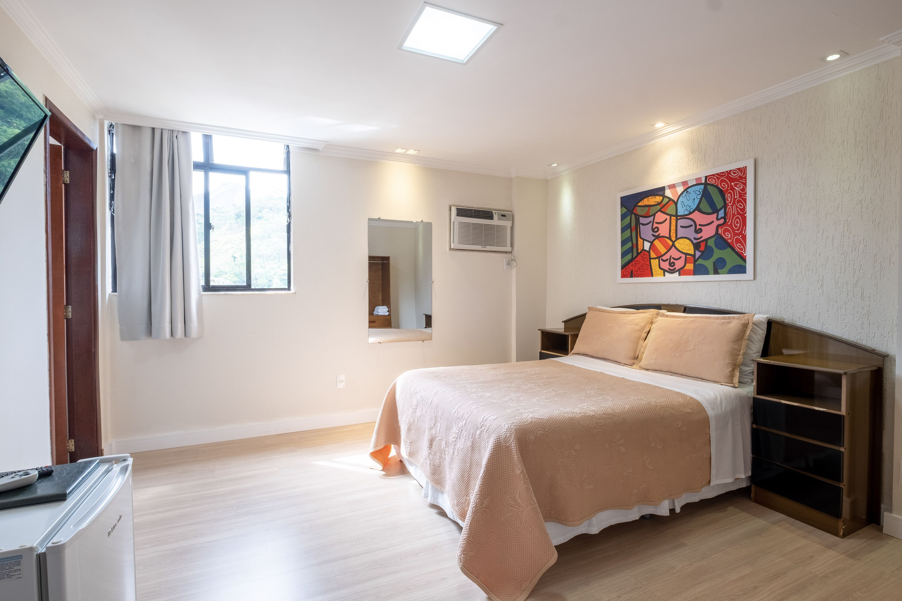 A bedroom with a bed, bedside tables, a window with curtains, an air conditioning unit, and a painting on the wall.