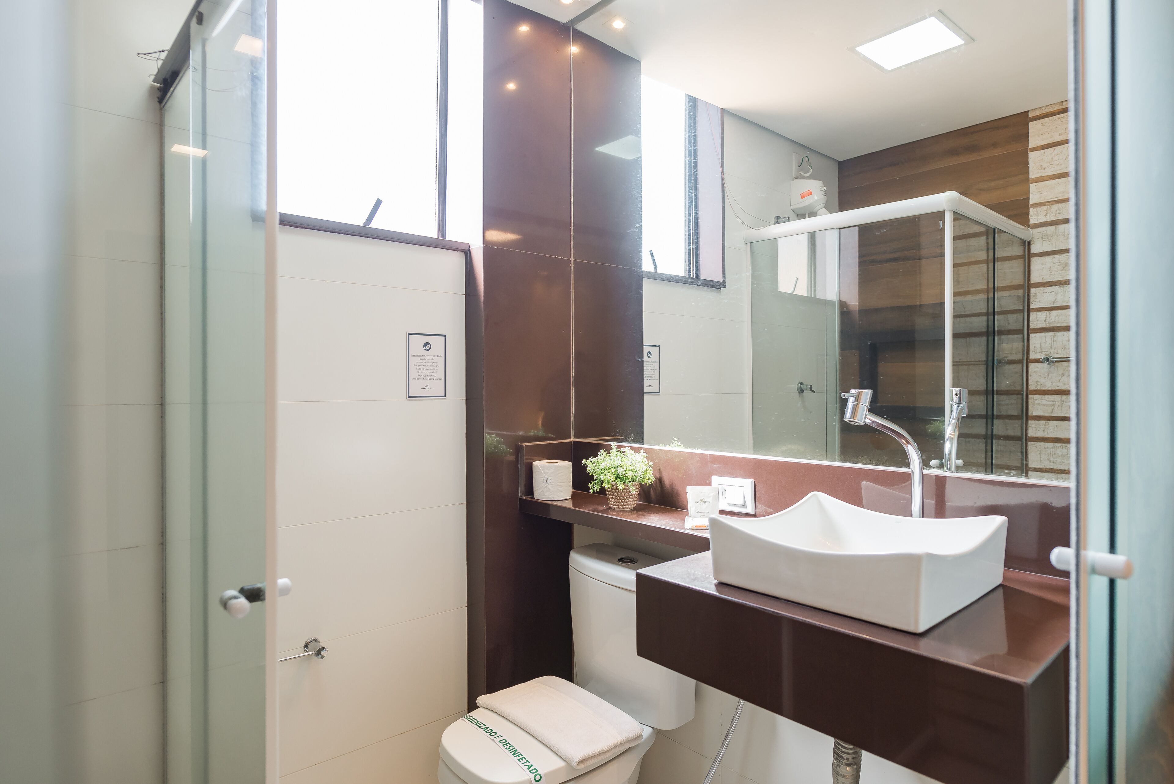 Un baño moderno con cabina de ducha de vidrio, inodoro blanco, tocador marrón con lavabo blanco y espejo sobre el lavabo.