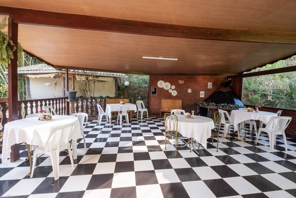 An outdoor patio with checkered flooring, white tables and chairs, a brick wall, and a grill area. - Hotel Serra Everest (Nova Friburgo)