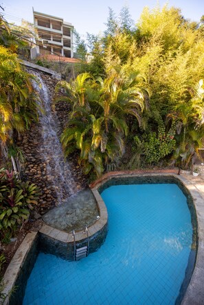 A swimming pool with a waterfall feature, surrounded by lush greenery and a multi-story building in the background. - Hotel Serra Everest (Nova Friburgo)