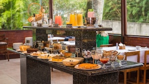 A buffet with various food items and drinks, including cakes, pies, and juices, displayed on a marble counter with a view of greenery outside.
