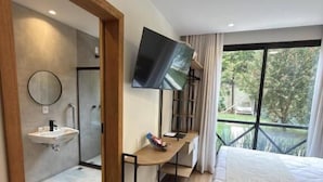 A hotel room with a bed, a TV mounted on the wall, a bedside table with a lamp, a mirror, and a window with a view of greenery.