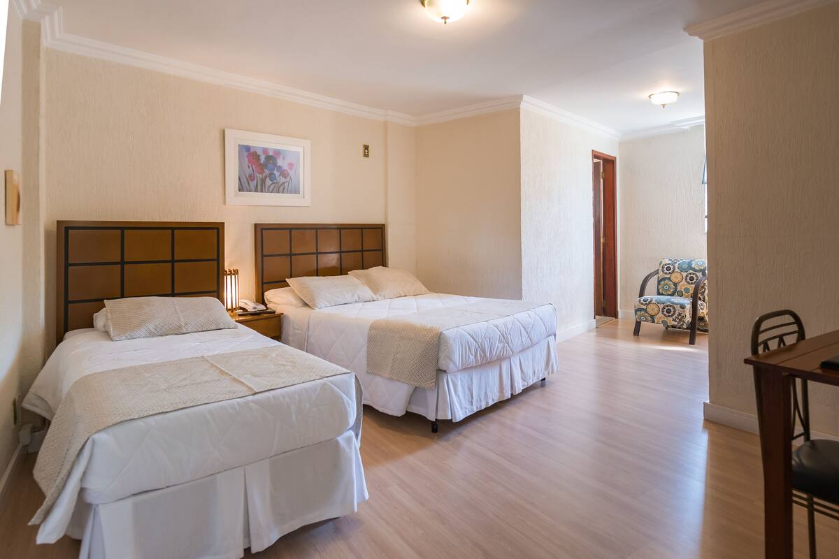 A hotel room with two beds, a wooden headboard, a bedside table with a lamp, and a chair with a patterned cushion.