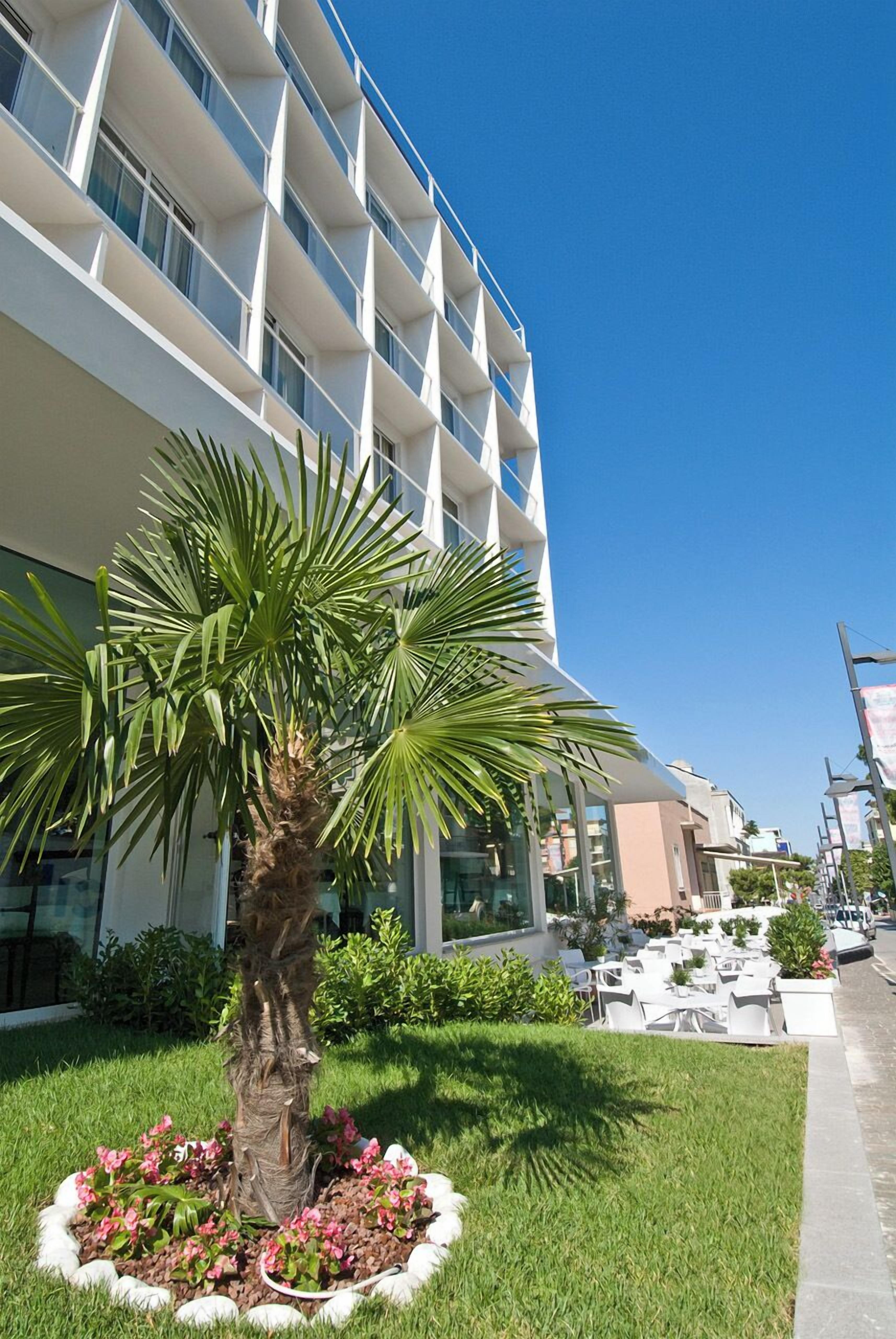 A modern hotel with a palm tree and flower bed in the foreground, and a pool area with outdoor seating in the background.