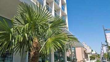 A modern hotel with a palm tree and flower bed in the foreground, and a pool area with outdoor seating in the background.