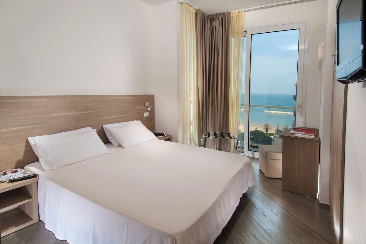 Habitación de hotel con una cama grande, una mesita de noche con teléfono y control remoto, un televisor montado en la pared y vista al mar a través de un amplio ventanal.
