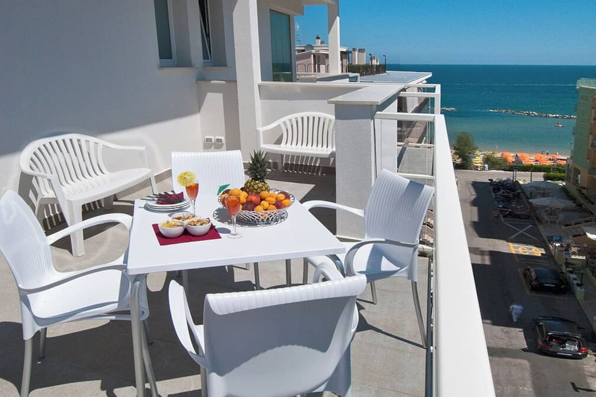 Un balcón con sillas blancas y una mesa dispuesta con frutas y una bebida, con vista a una playa y al océano.