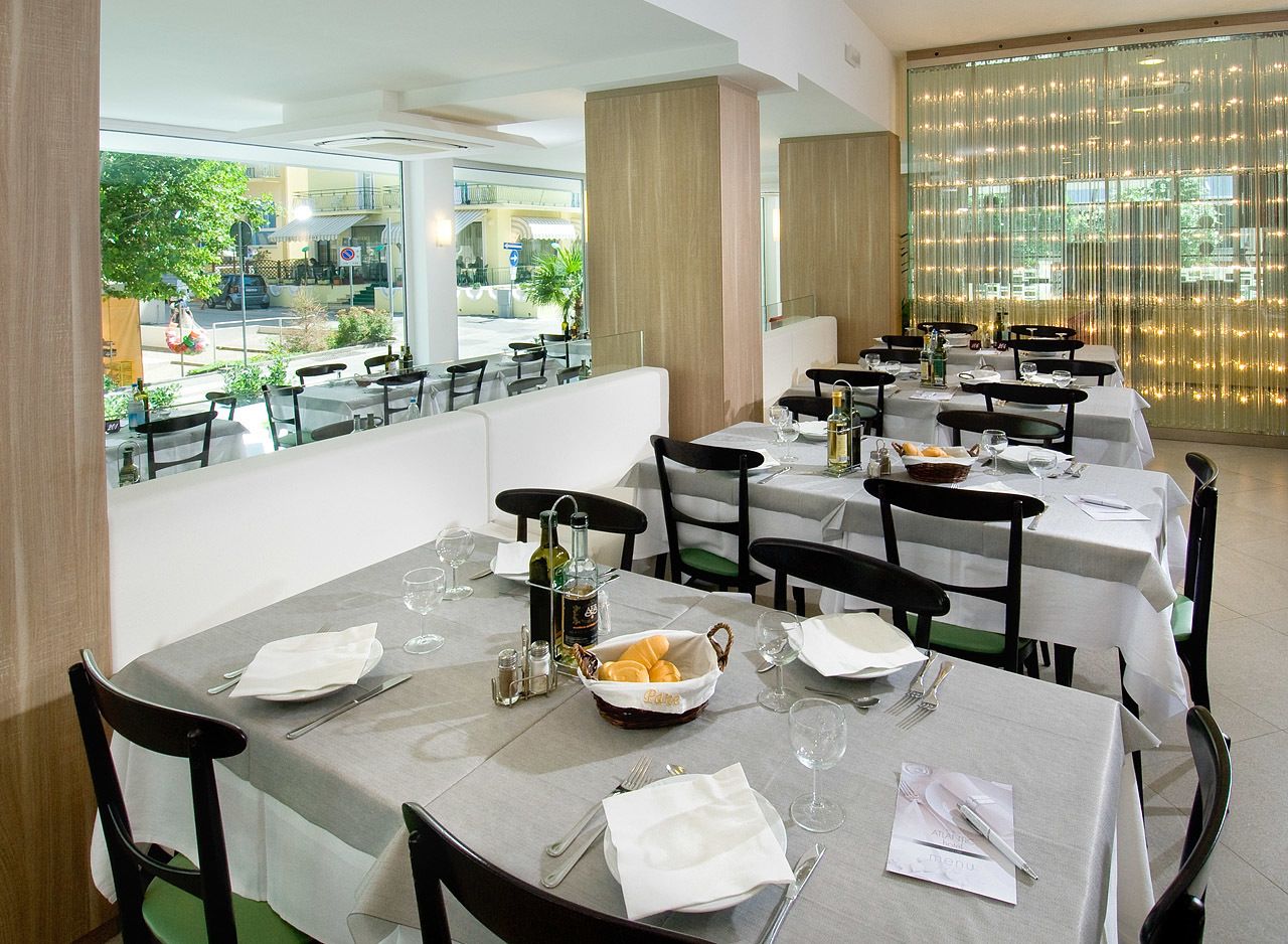 A restaurant with multiple tables set for dining, featuring a view of the street and greenery outside.