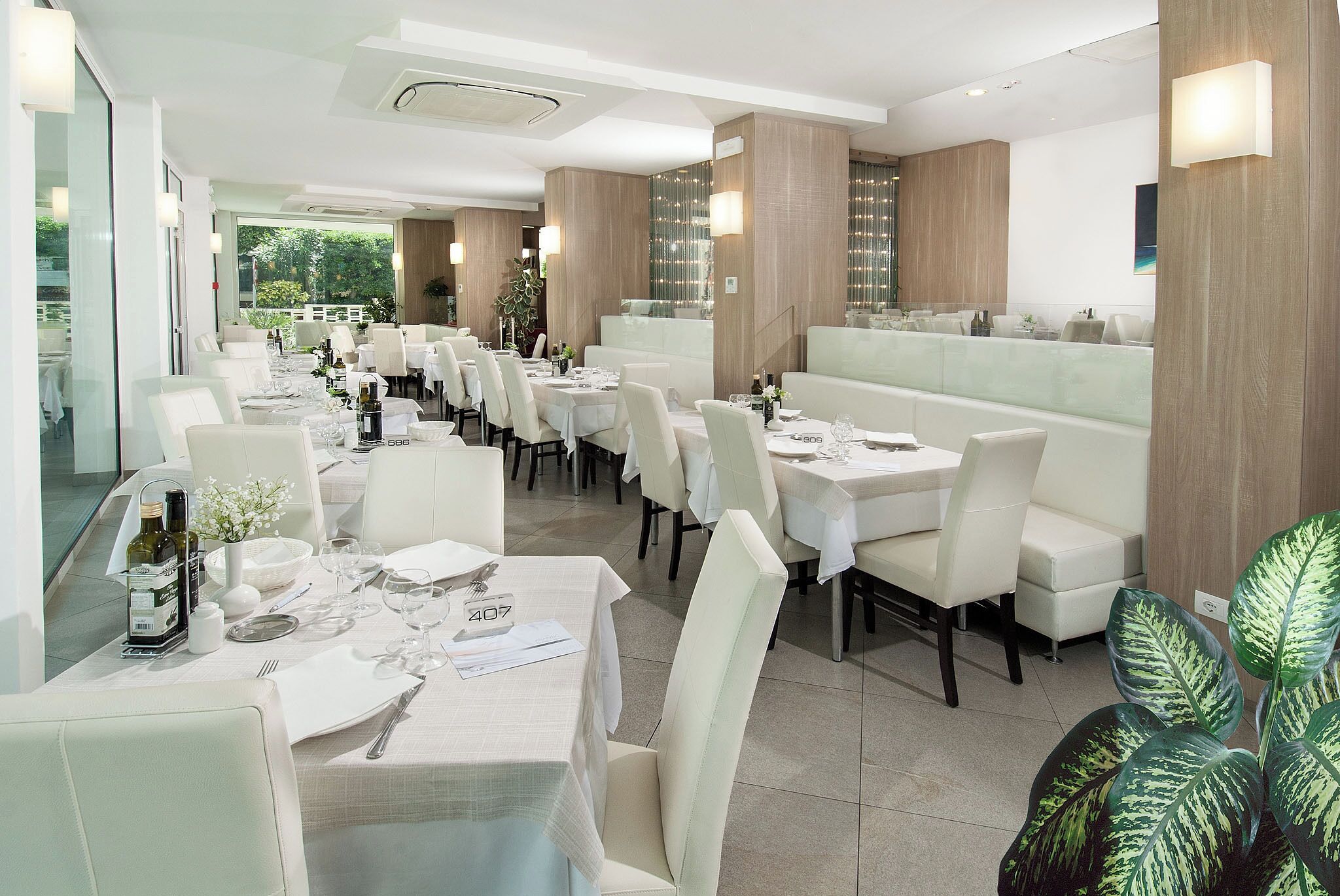 A modern restaurant with white tables and chairs, a bar area, and a view of greenery outside.