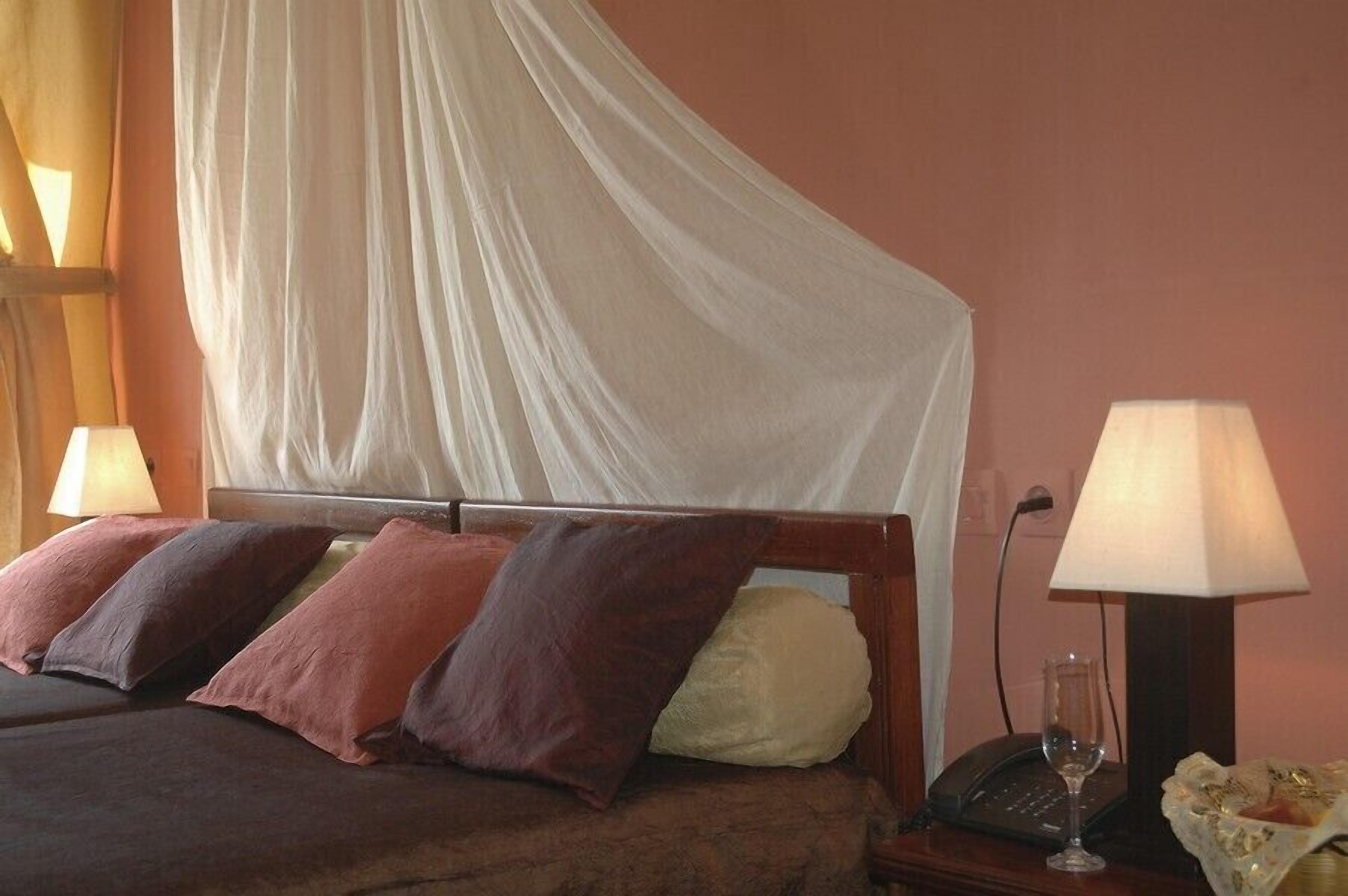 Een bed met baldakijn, meerdere kussens, een bedlampje, een telefoon en een glas wijn op een nachtkastje van hout.