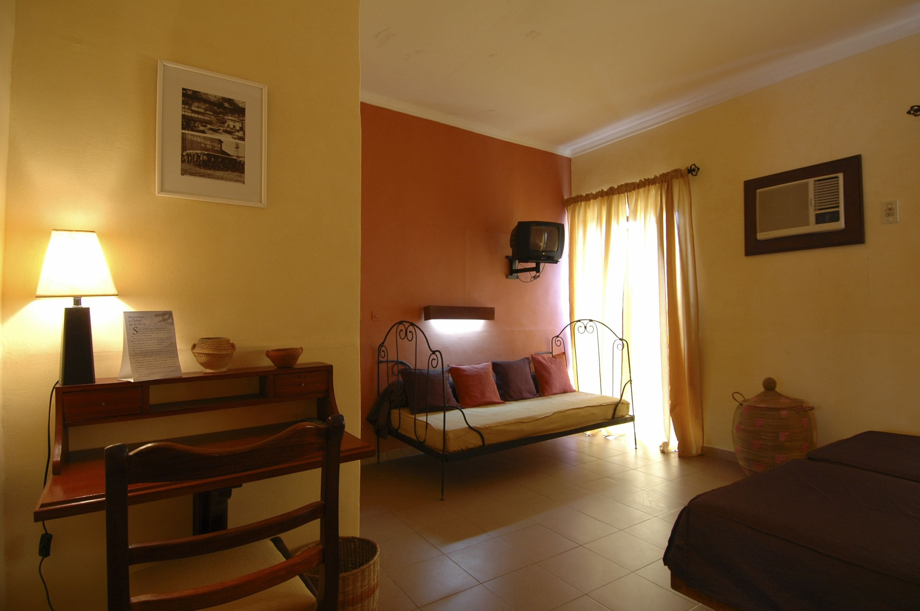 Una habitación con cama, escritorio con lámpara, silla, televisor montado en la pared y ventana con cortinas.