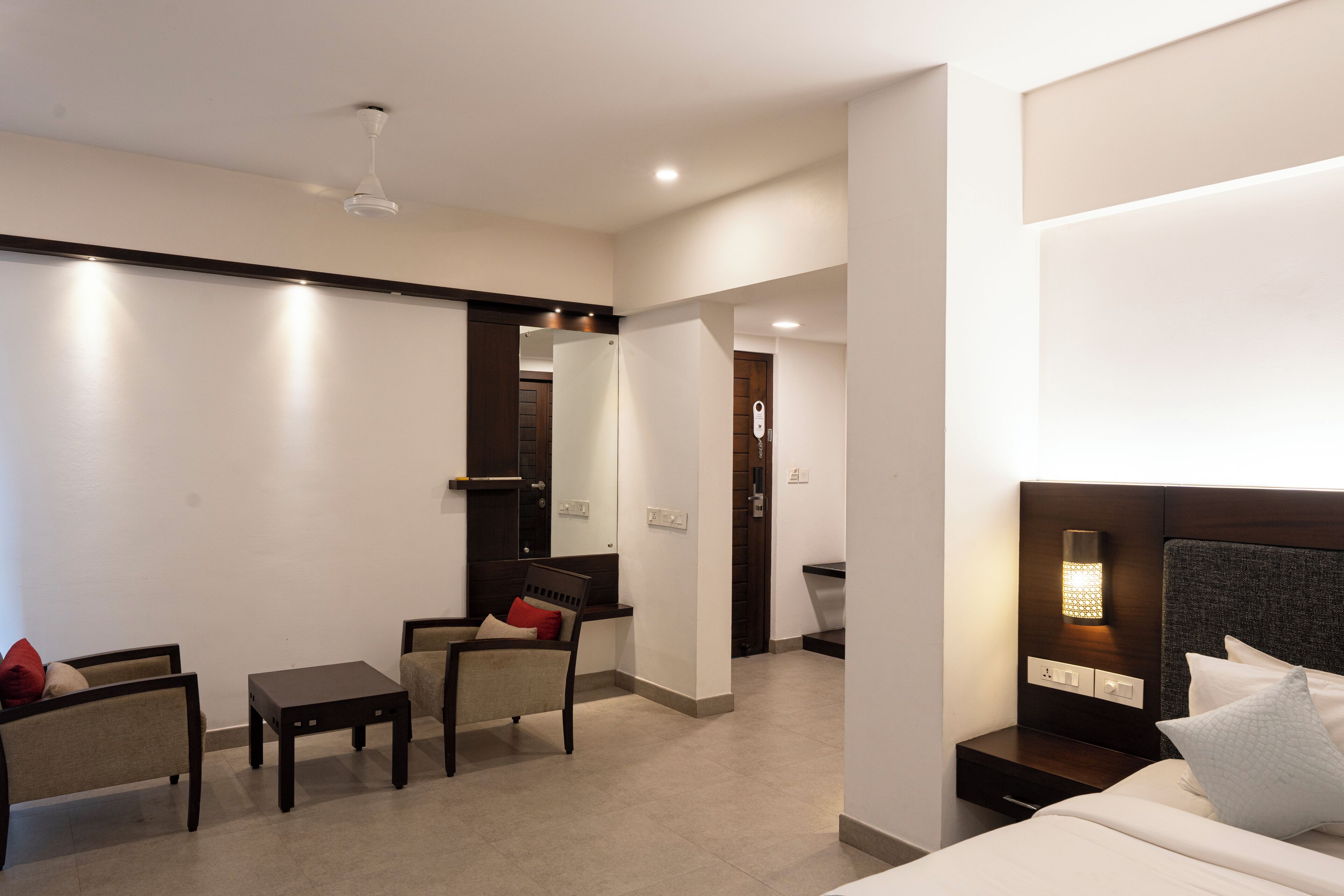A modern hotel room with a bed, bedside table, and a small desk with a chair. There is a mirror on the wall and a light fixture above the desk.