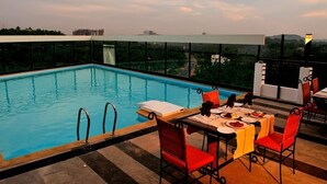 A rooftop pool with a dining area, surrounded by a glass barrier, offering a view of the cityscape. - Copper Folia (Kozhikode)