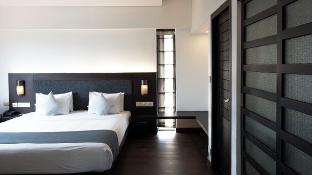 A modern hotel room with a large bed, a nightstand, a desk, and a view of the city through a window. - Copper Folia (Kozhikode)