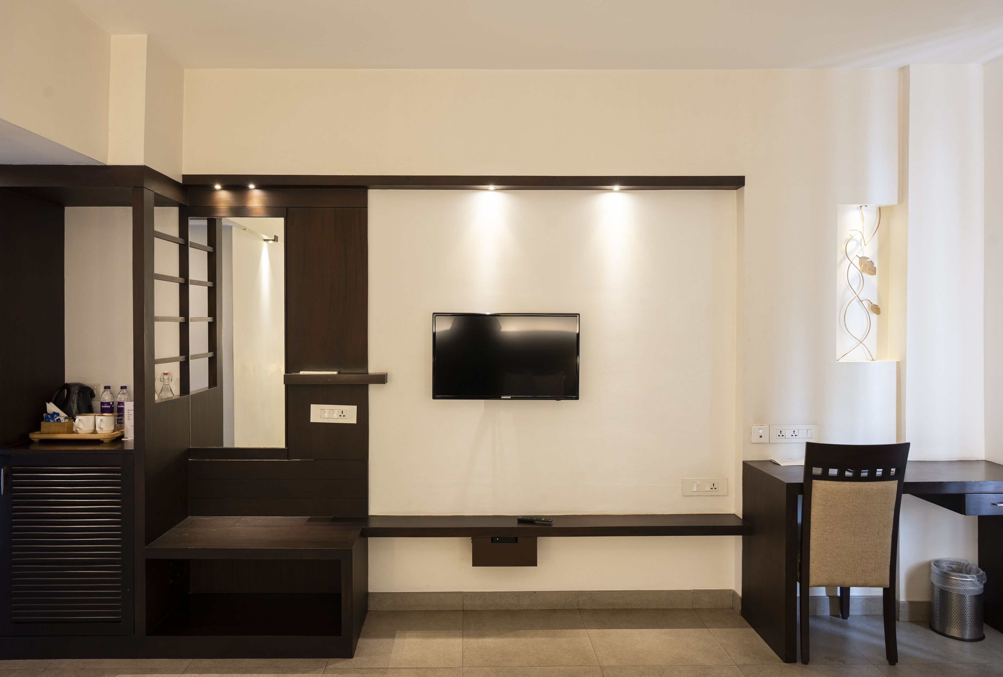A modern hotel room with a flat-screen TV mounted on the wall, a small desk with a chair, a bench, and a trash can.