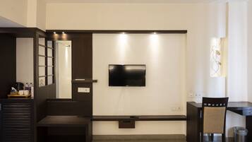 A modern hotel room with a flat-screen TV mounted on the wall, a small desk with a chair, a bench, and a trash can.
