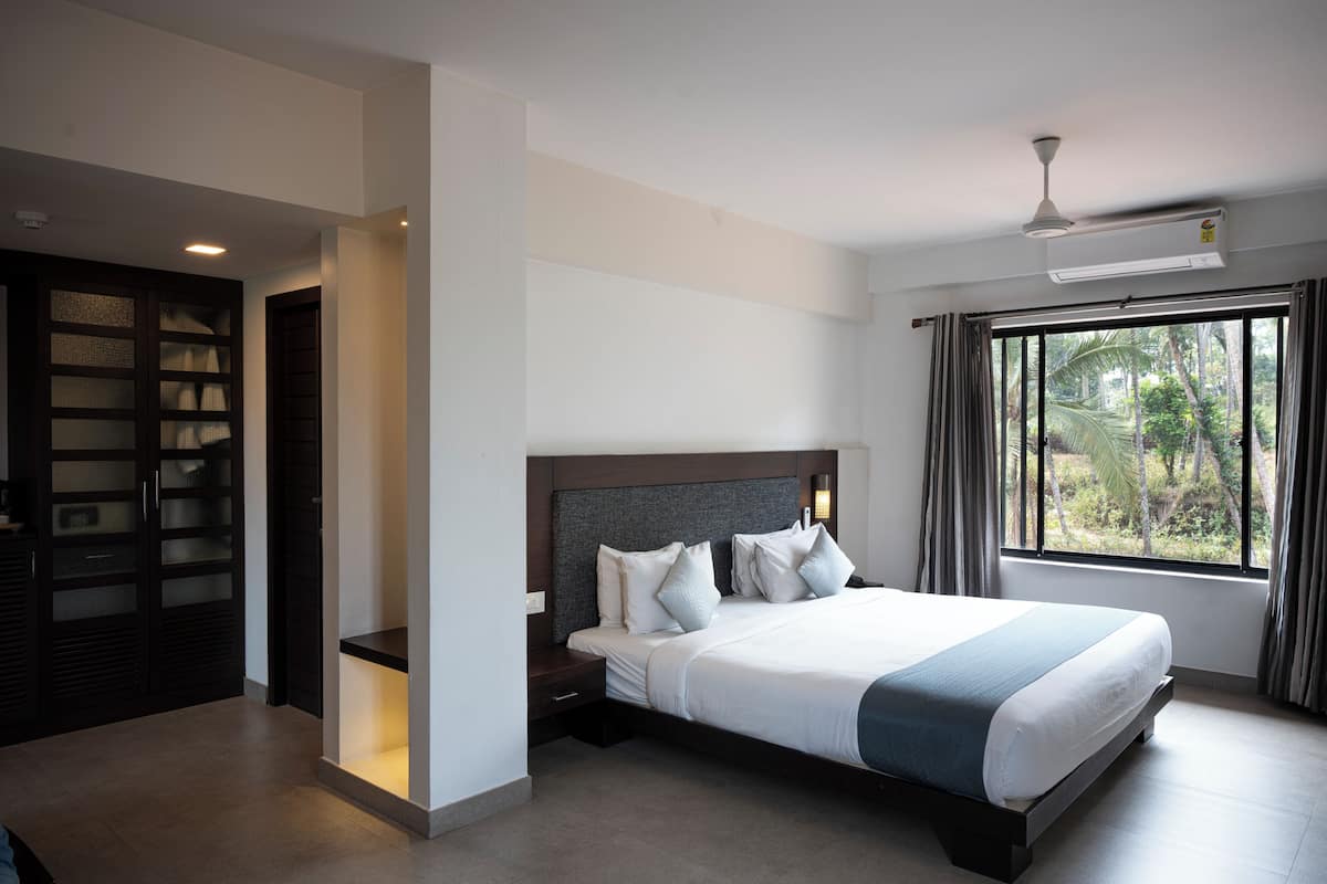 A modern hotel room with a large bed, a ceiling fan, an air conditioning unit, a window with curtains, and a built-in wardrobe.