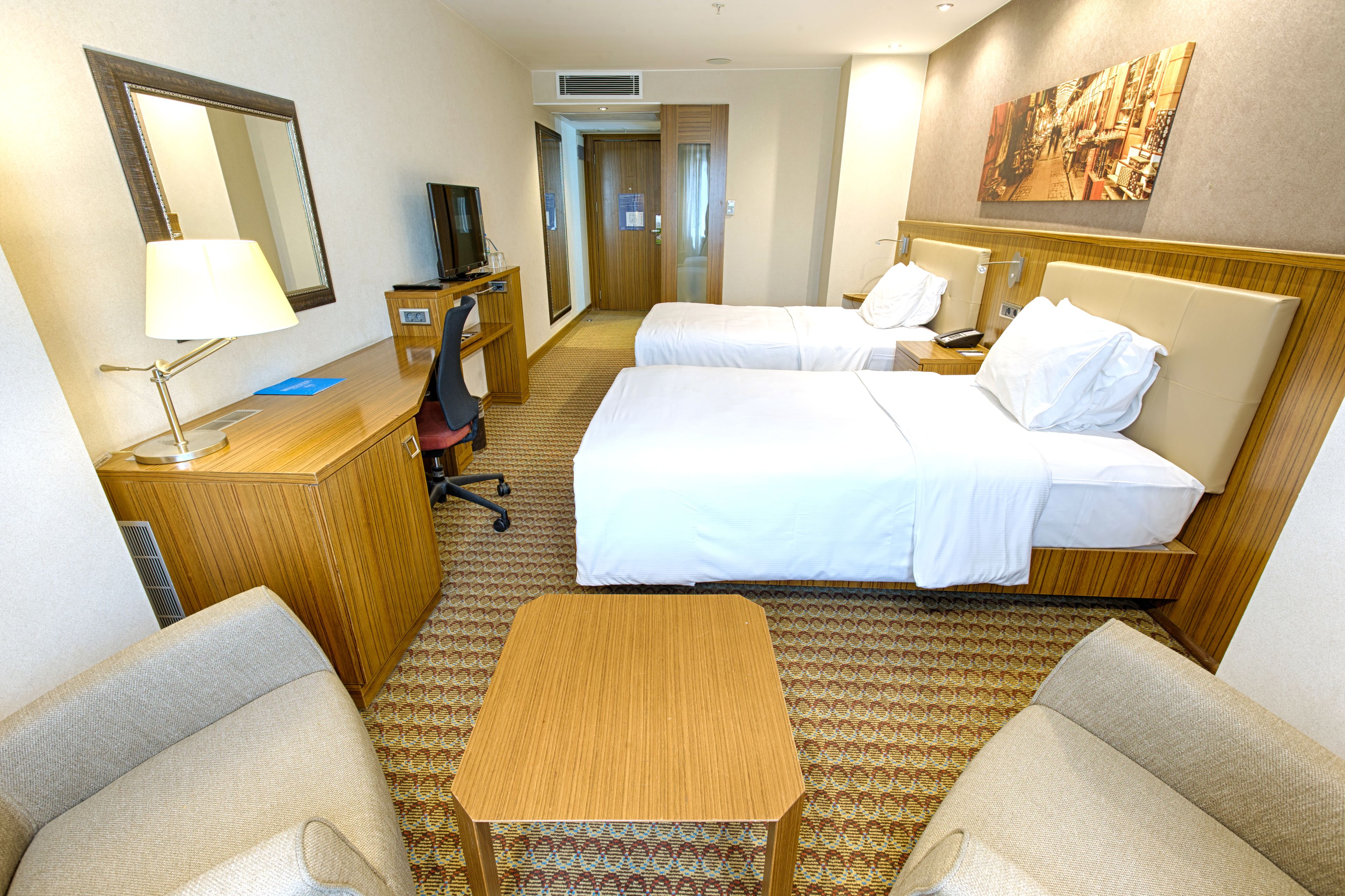 A hotel room with two beds, a desk, a chair, a TV, and a painting on the wall.