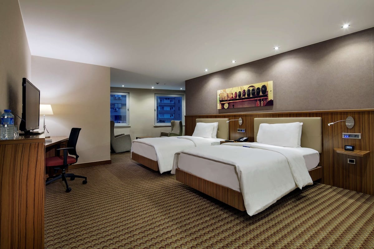A hotel room with two beds, a desk with a chair, a TV, and a wall-mounted shelf with decorative items.