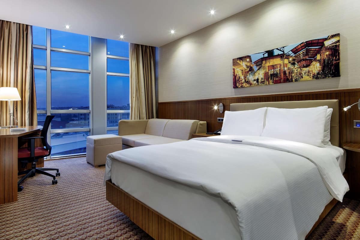 A hotel room with a large bed, a desk with a computer, a chair, and a view of the city through large windows.