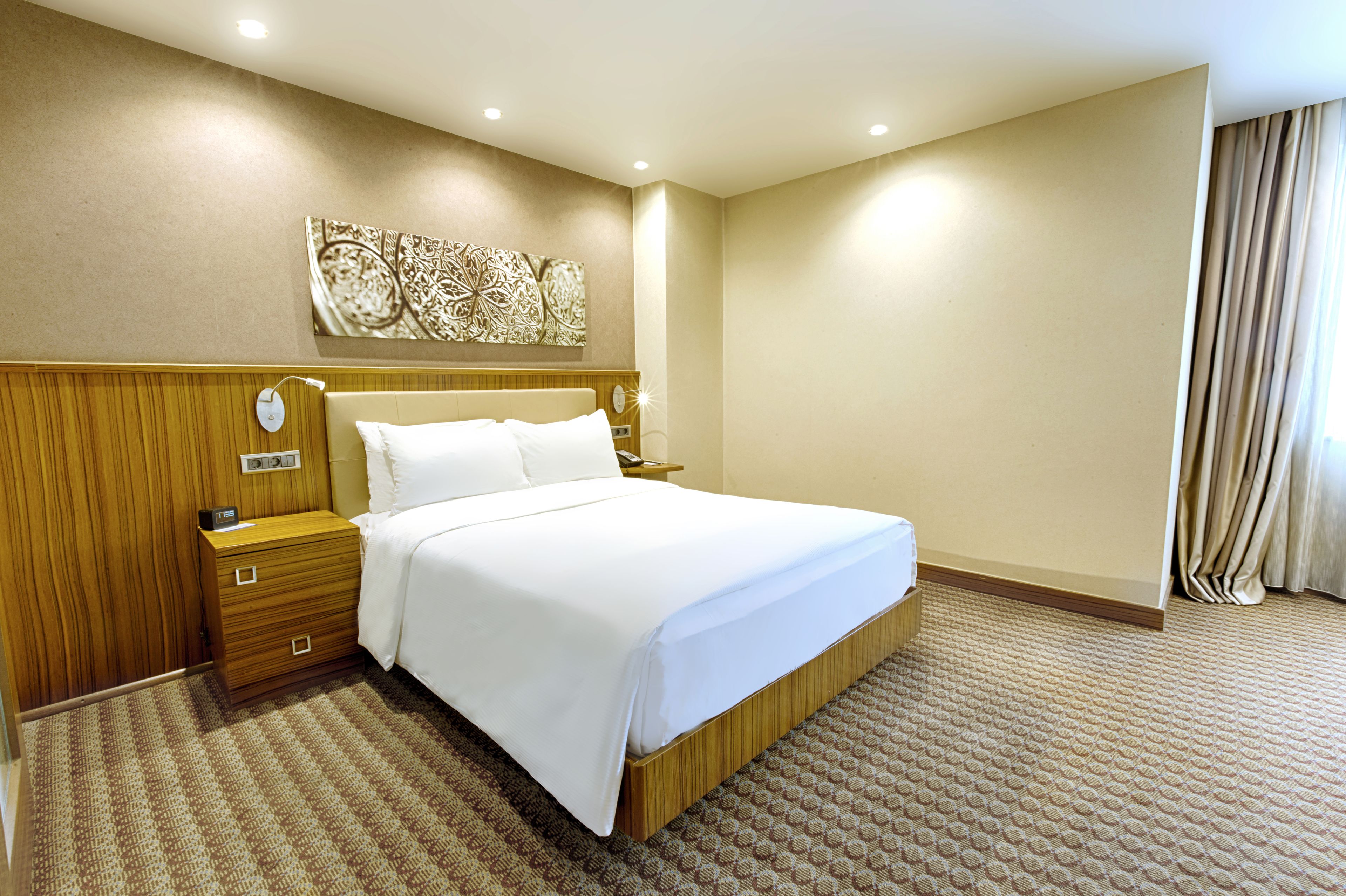 Habitación de hotel con una cama grande, cabecera de madera, mesitas de noche, un reloj de pared y una alfombra estampada.