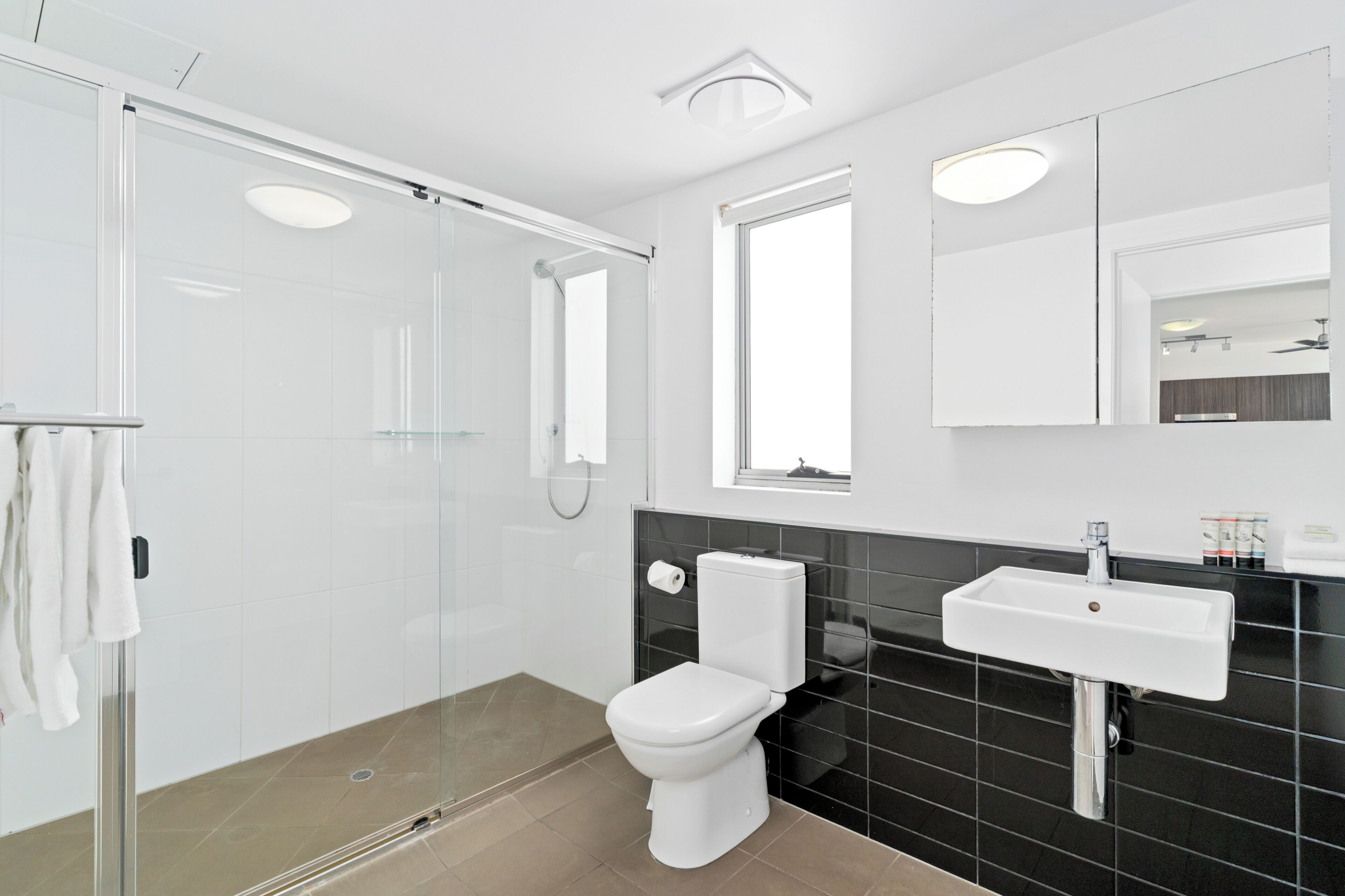 A modern bathroom with a glass shower enclosure, a white toilet, a black tiled backsplash, and a white sink.