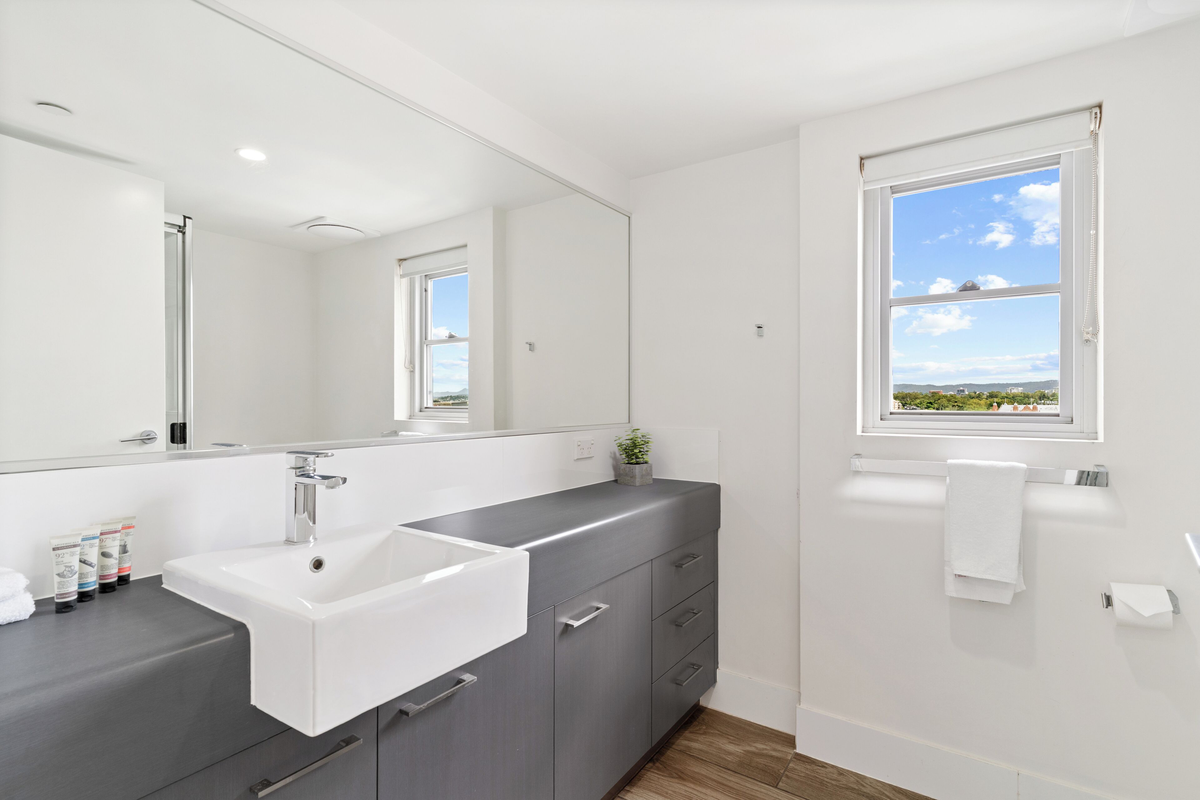 Un baño moderno con un espejo grande, un lavabo blanco, armarios grises, una ventana con vistas y un perchero para toallas.