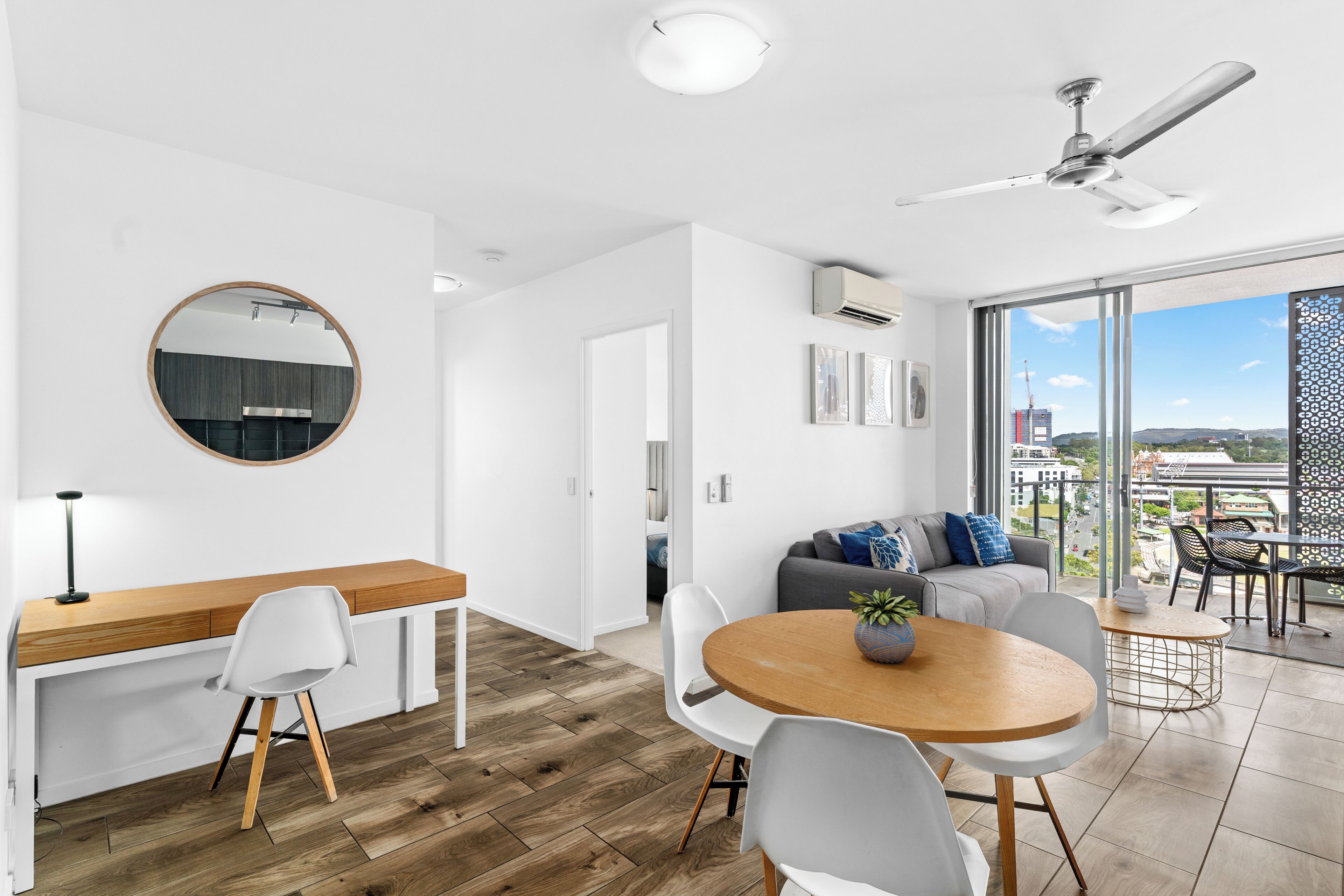 Un espacio habitable moderno con una mesa de comedor de madera, sillas blancas, un sofá gris con cojines azules, un espejo redondo y un ventilador de techo.