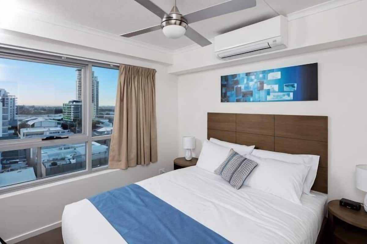Una habitación de hotel con una cama grande, un ventilador de techo, una ventana con vista a edificios y un cuadro en la pared.