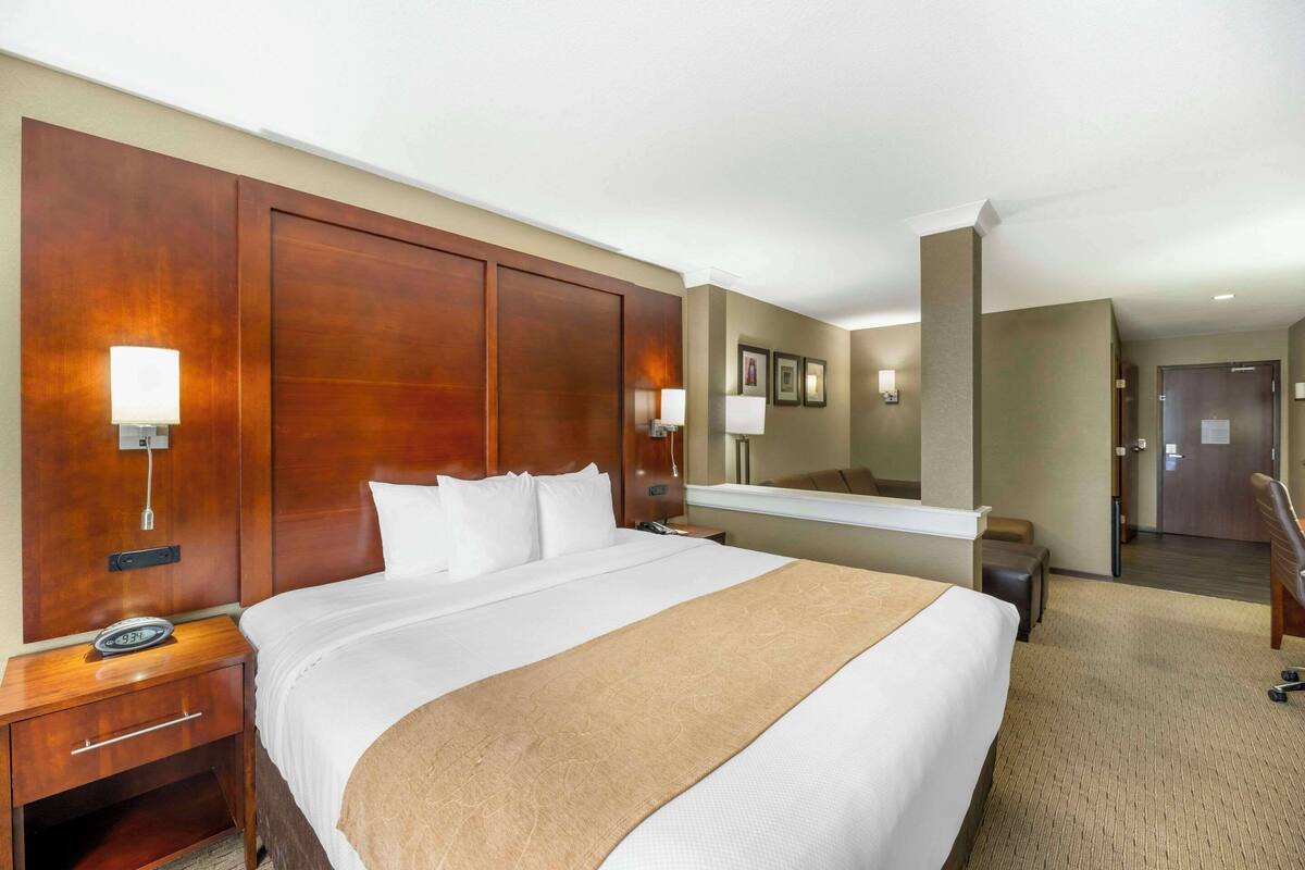 A hotel room with a large bed, a wooden headboard, bedside lamps, a nightstand, a desk, a mirror, a chair, and a door.