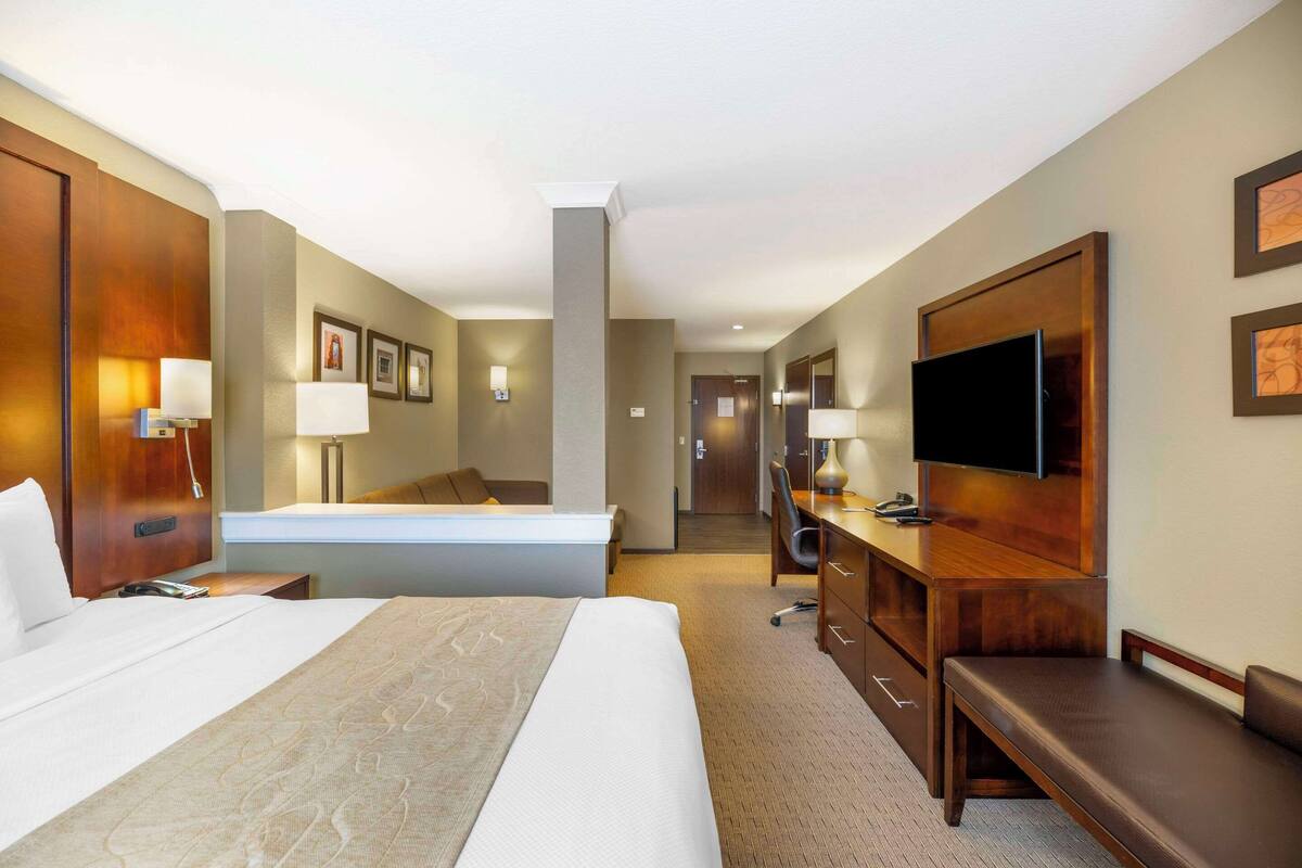 A hotel room with a large bed, a desk with a flat-screen TV, a chair, a desk lamp, and a wall-mounted TV.