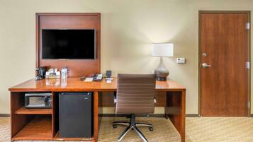 A hotel room with a wooden desk, a television, a microwave, a refrigerator, a lamp, and a door.