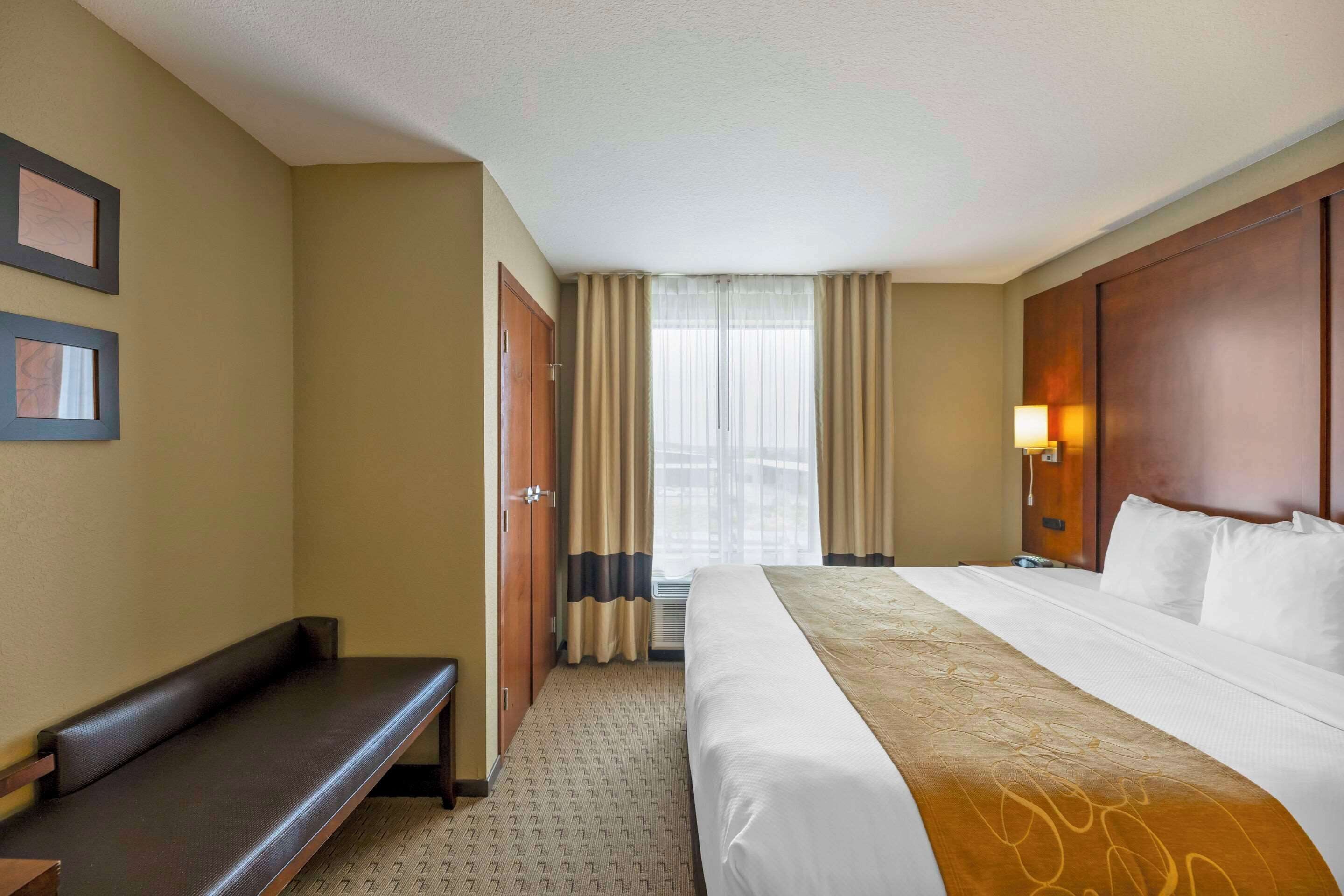 A hotel room with a large bed, a bench, a desk, and a window with curtains.