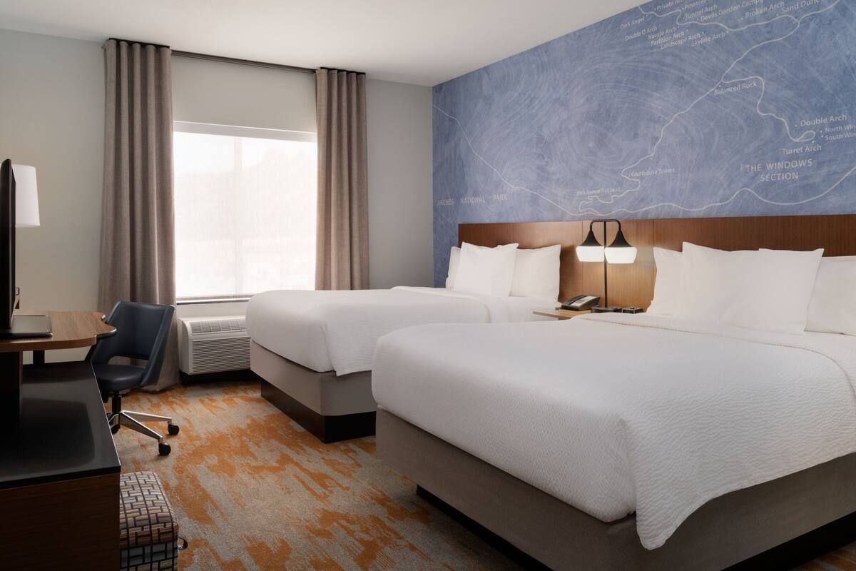 A hotel room with two beds, a desk, a chair, and a large window with curtains.