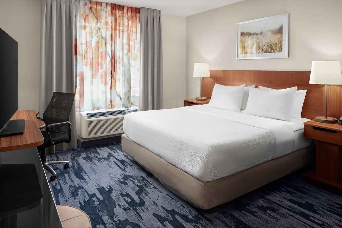 A hotel room with a large bed, two bedside tables with lamps, a desk with a chair, an air conditioning unit, and a window with curtains.