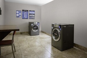 A laundry room with two front-loading washing machines, a table, and informational posters on the wall. - Hilton Garden Inn Denison/Sherman/At Texoma Event Center (Denison)