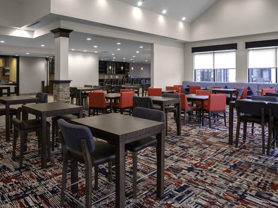 Espaço de refeições de hotel com várias mesas e cadeiras, cozinha ao fundo e grandes janelas que proporcionam luz natural.