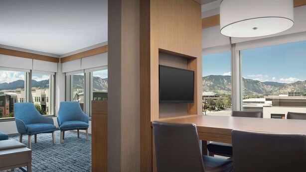 A modern hotel room with a large window, a flat-screen TV mounted on a wooden panel, and a dining area with a table and chairs.