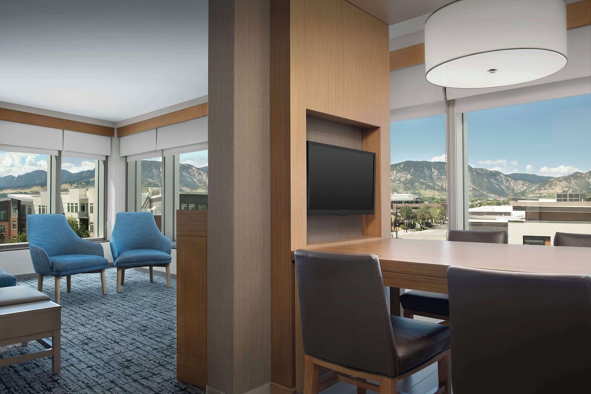 A modern hotel room with a large window, a flat-screen TV mounted on a wooden panel, and a dining area with a table and chairs.
