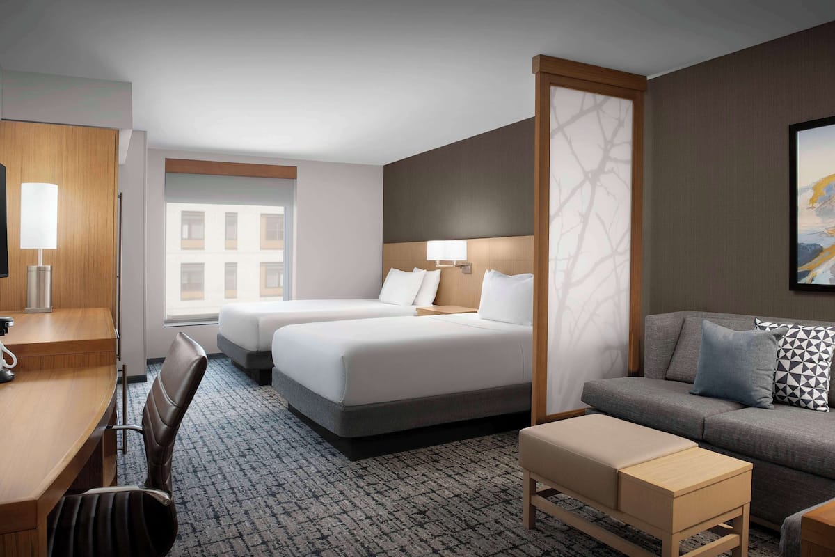 A hotel room with two beds, a desk, a sofa, and a large window.