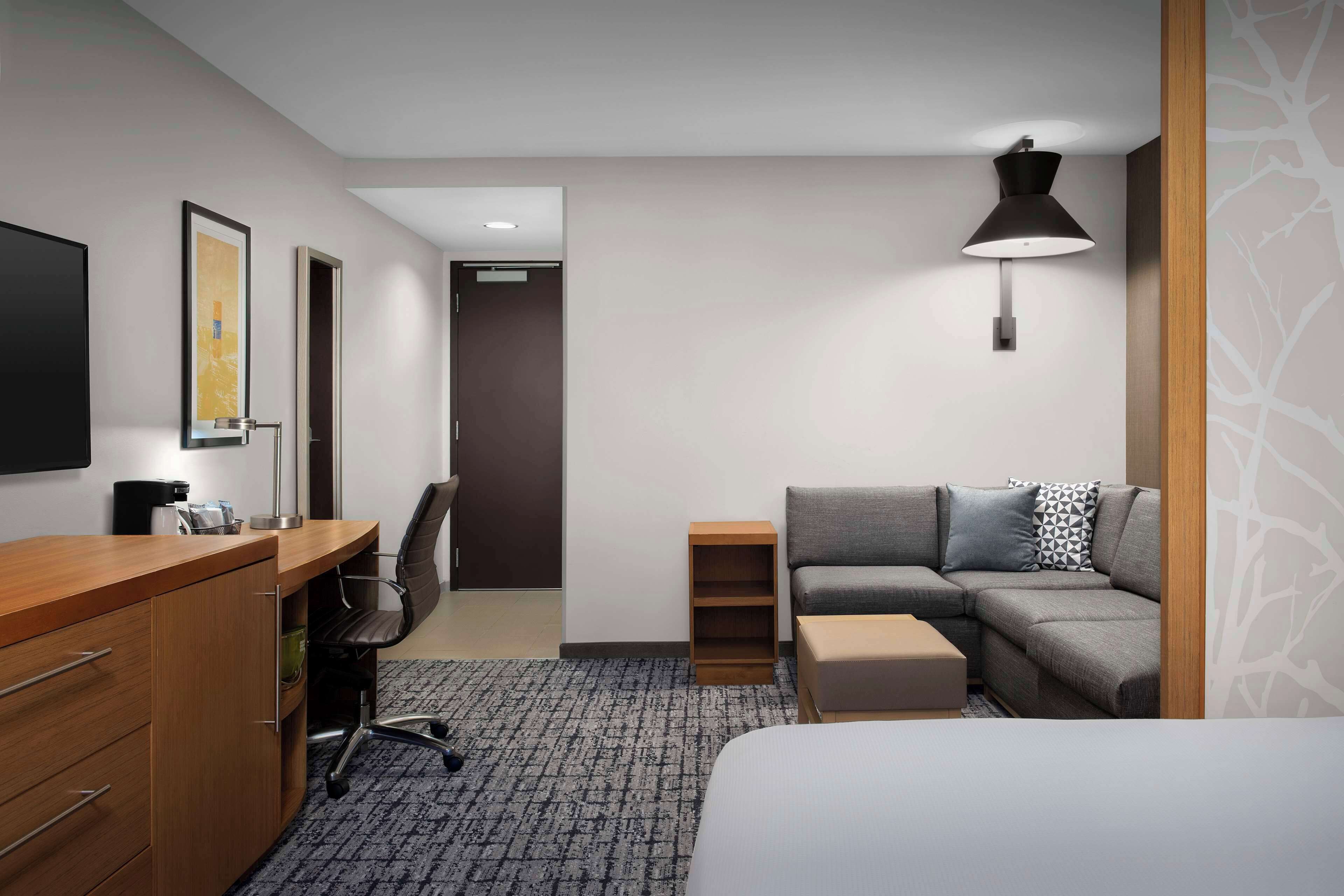 Habitación de hotel con un escritorio de madera, un sofá gris, una cama, una lámpara de pie y un cuadro en la pared.