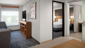 A hotel room with a large window, a TV on a wooden stand, a bed with white linens, and a mirror reflecting the room.