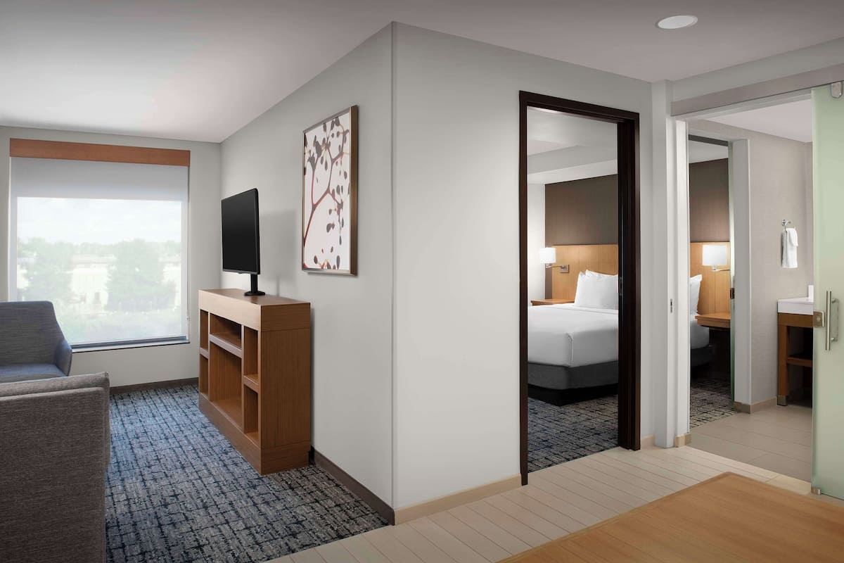 A hotel room with a large window, a TV on a wooden stand, a bed with white linens, and a mirror reflecting the room.