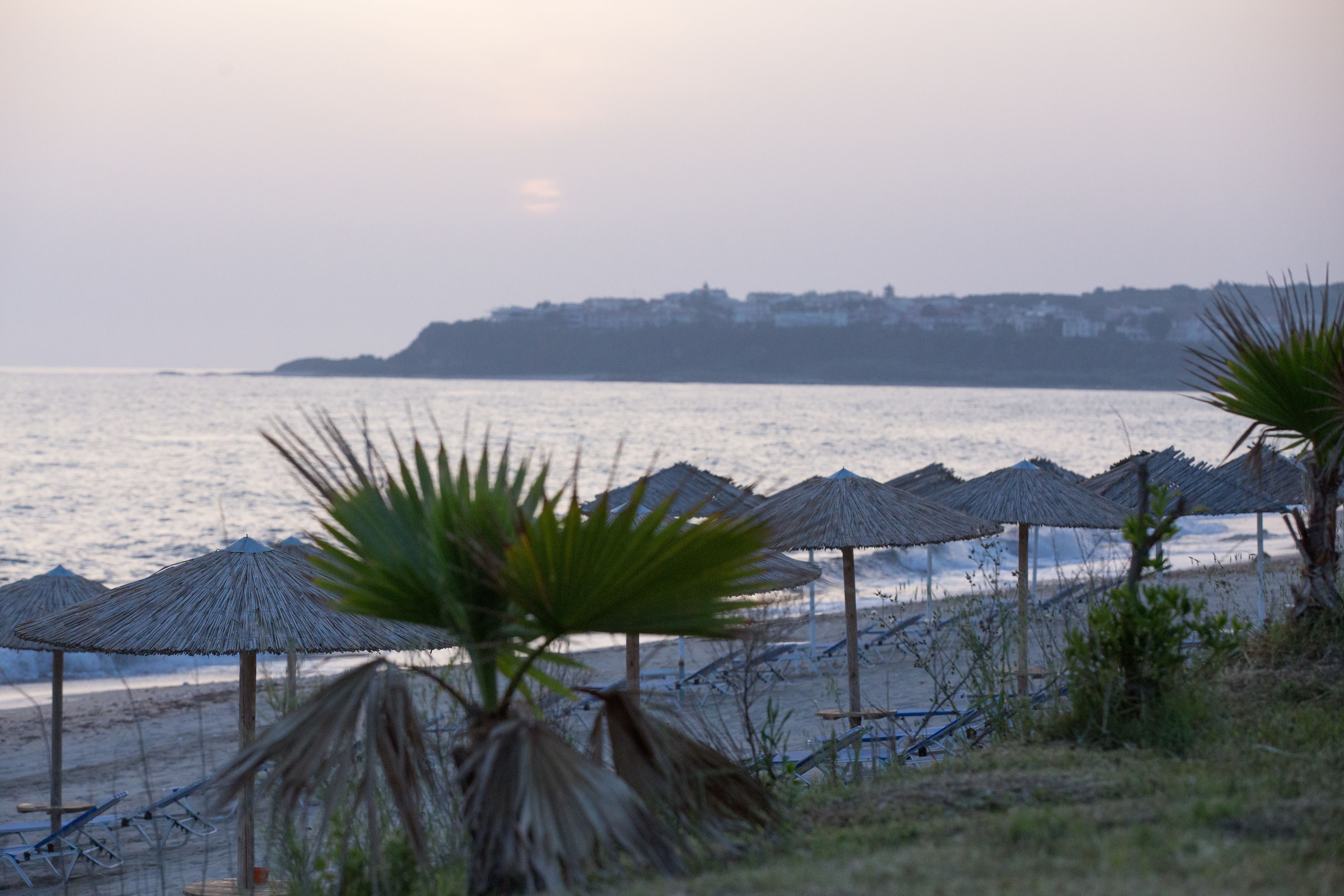 A beach with thatched umbrellas and lounge chairs, a calm sea, and a distant hill with buildings.
