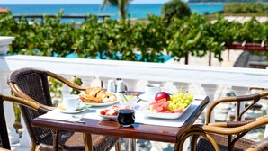 A dining table with breakfast items, a glass of red wine, and a view of the ocean and vineyards. - Poseidon Beach Hotel (Preveza)