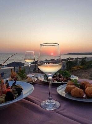 A table set with plates of seafood and croquettes, two wine glasses, and a sunset view. - Poseidon Beach Hotel (Preveza)