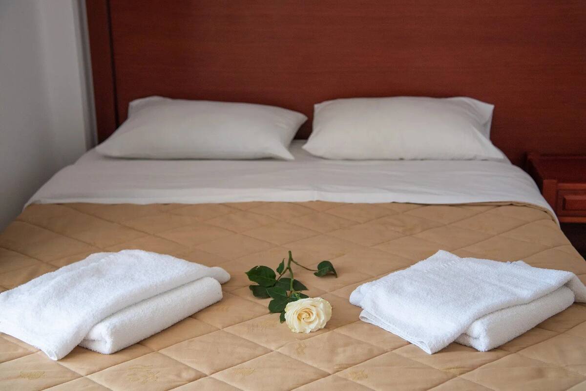 A neatly made bed with white linens, two pillows, and two folded towels on a beige quilted bedspread. A white rose is placed in the center.