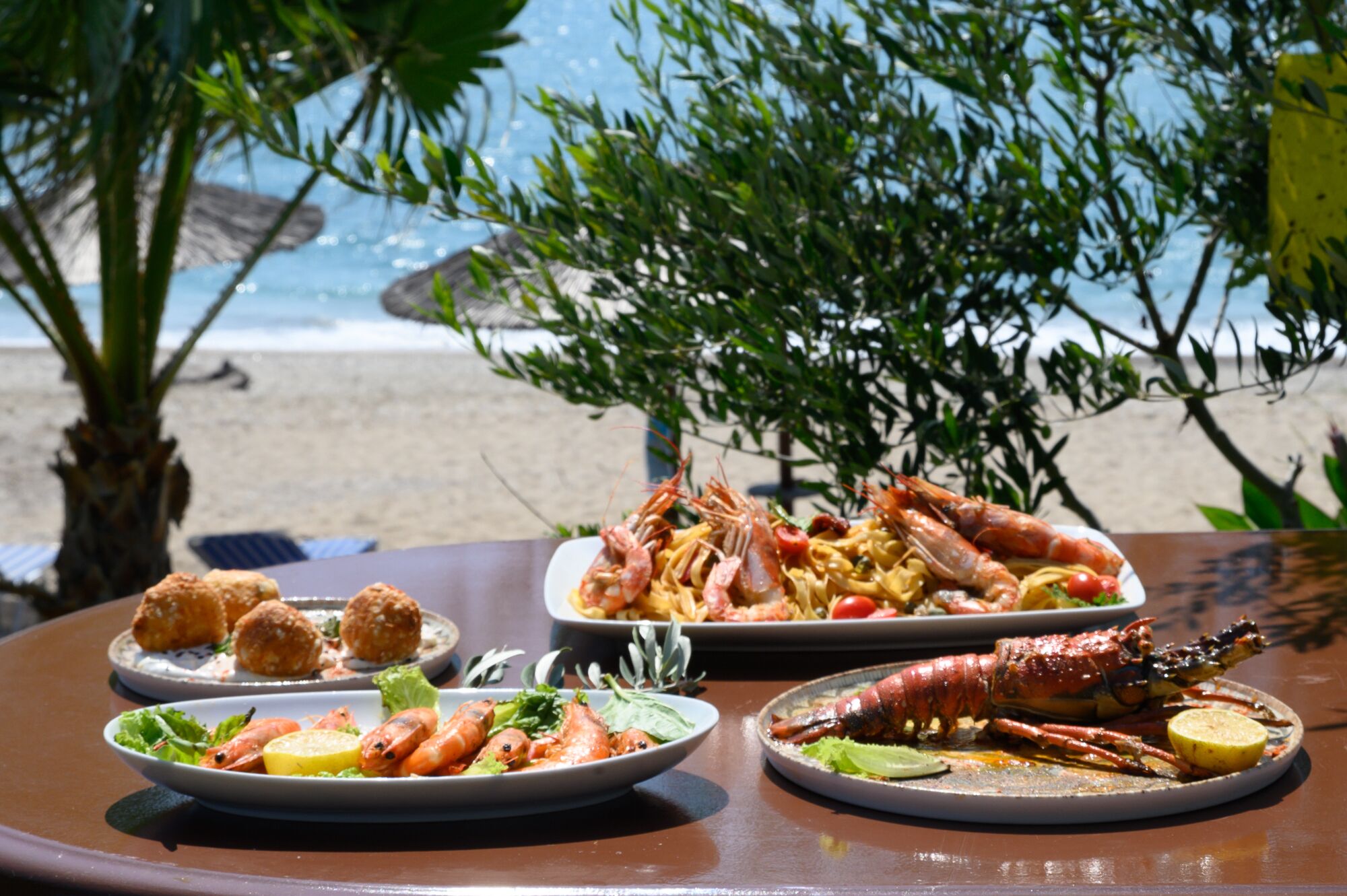 Un tavolo imbandito con piatti di pesce, tra cui gamberi e aragosta, su una spiaggia con palme e l'oceano sullo sfondo.
