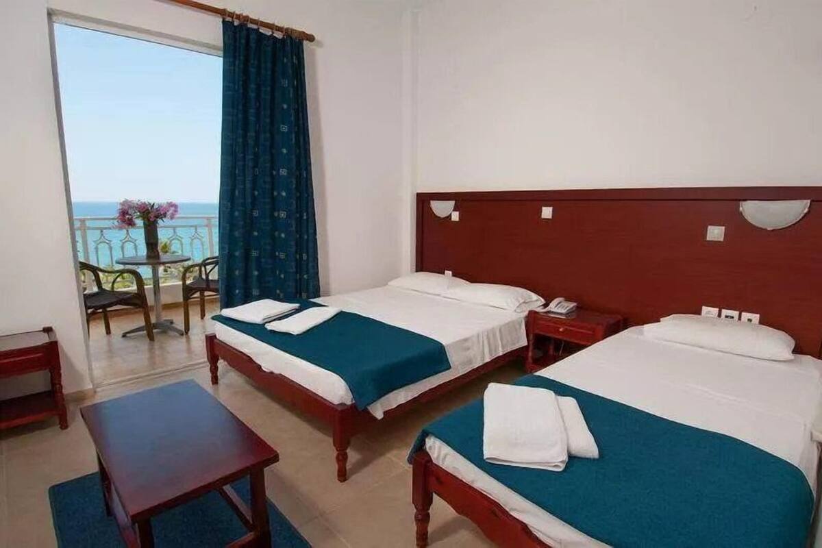 A hotel room with two beds, a small table, and a view of the sea through a balcony.